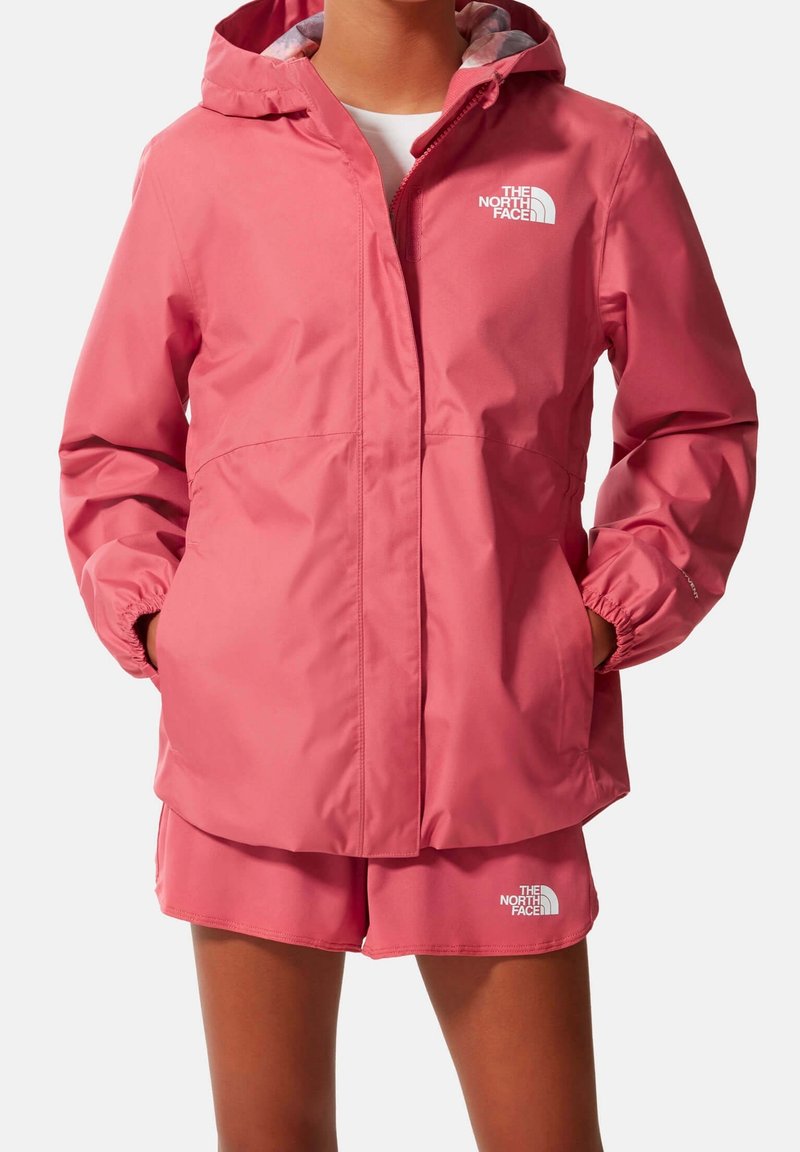 Person som bär en rosa North Face-huvjacka och matchande shorts, händerna i jackfickorna, vit bakgrund.