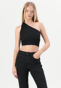 Joven mujer con largo cabello rubio, vistiendo un top corto negro de un solo hombro y jeans negros de alta cintura, con la mano en el bolsillo, frente a un fondo blanco.