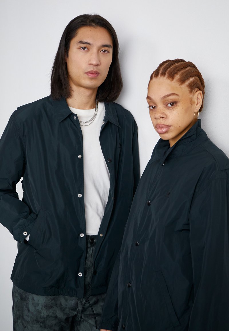 Due modelli indossano giacche leggere di un elegante blu navy, realizzate in tessuto liscio, con bottoni a pressione e colletti standard.