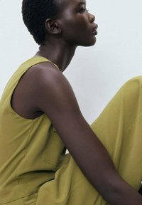 Olive green sleeveless dress with a relaxed fit, smooth texture, and a V-neckline, showcased on a model in profile against a neutral background.