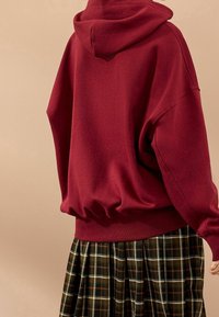 Donkerrode oversized hoodie met een capuchon en trekkoord, geribbelde manchetten en een geplooide zoom, gecombineerd met een geruite rok in donker groene en bruine tinten.