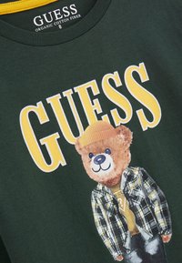 Tricou de culoare verde închis cu un personaj urs în cămașă în carouri și cu o căciulă galbenă. Textul din față "GUESS" în litere galbene îngroșate. Material din bumbac organic.