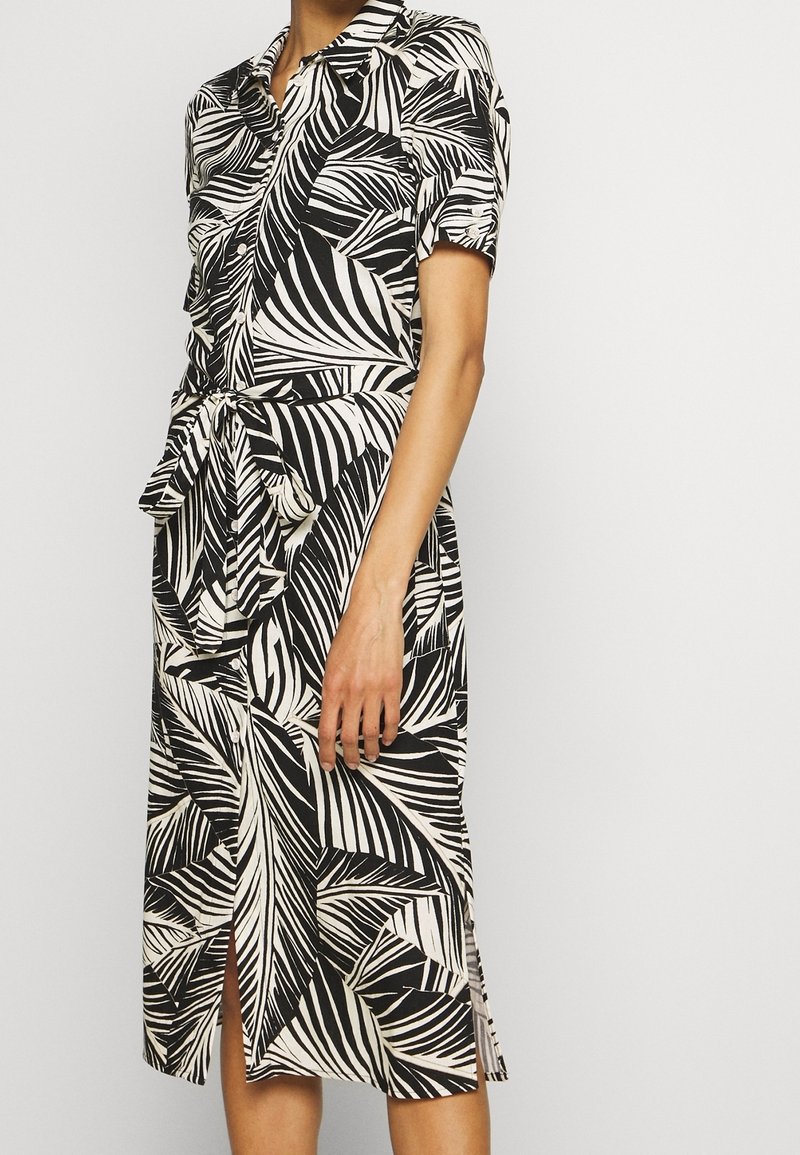 Femme portant une robe chemise imprimé feuilles tropicales noir et blanc, manches courtes et ceinture nouée à la taille.