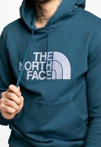 Tyrkysová bavlněná mikina s kapucí, s velkým bílým logem "THE NORTH FACE" na hrudi, se stahovací šňůrkou na kapuci a přední klokankovou kapsou.