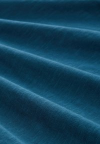 Tessuto in un profondo teal, che mostra una superficie morbida e testurizzata con pieghe orizzontali distintive e una finitura liscia, indicando un materiale leggero.