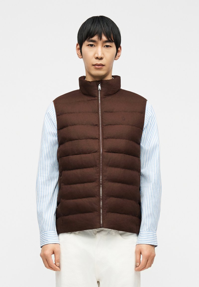 Άνδρας που φοράει καφέ αμάνικο φουσκωτό γιλέκο πάνω από μπλε και λευκή ριγέ μπλούζα με λευκό παντελόνι, στέκεται μπροστά σε απλό ανοιχτόχρωμο φόντο.