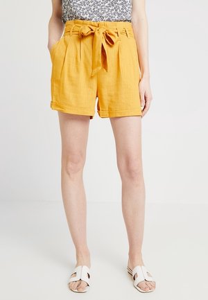 Femme portant un short taille haute moutarde noué avec des revers retroussés, un haut imprimé et des sandales blanches à enfiler, debout devant un fond uni.