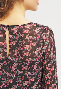 Blouse noire en tissu transparent avec des motifs floraux vibrants en rose, jaune et sarcelle. Dotée d'un dos avec ouverture en forme de goutte et de manches longues.