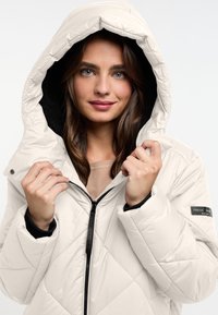 Pufferjack in lichtbeige met een diamanten gestikt patroon, voorzien van een capuchon en rits van zwarte fleece, met een opvallend logopatch op de mouw.
