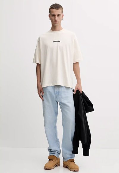 T-shirt crema con logo nero "BARROW", vestibilità rilassata, abbinata a jeans azzurri e calzature marroni. Giacca nera tenuta nella mano sinistra.