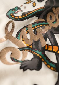 T-shirt grafico con lettering dorato testurizzato e disegno intricato di un serpente in verde e nero, su uno sfondo di tessuto crema chiaro.