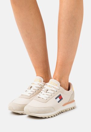 Sneaker beige con una combinazione di tessuto morbido e suede, con lacci bianchi e una patch logo in rosso, bianco e blu sul lato.