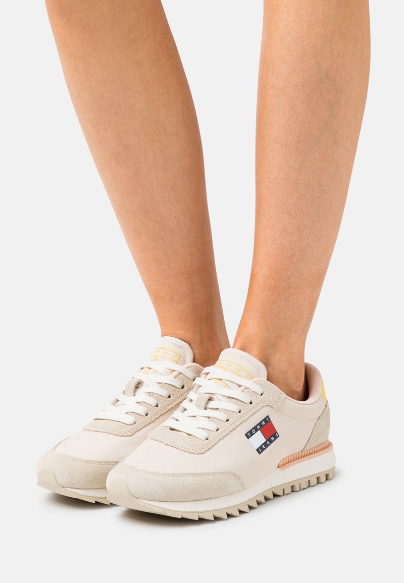 Beige sneakers med en kombination av slät tyg och mocka, med vita snören och en logotyp på sidan i röd, vit och blå.