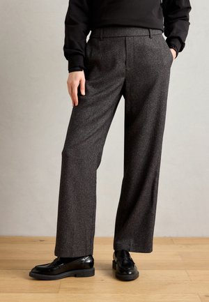 Osoba ubrana w ciemnoszare szerokie spodnie, z rękami w kieszeniach i czarne skórzane mokasyny, stojąca na drewnianej podłodze.