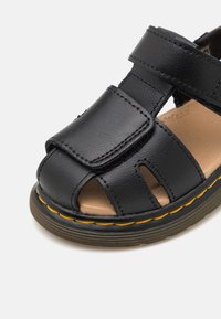 Sandal in pelle nera con design aperto sulla punta, cinturino in velcro regolabile e soletta marrone chiaro. Presenta cuciture gialle lungo la suola.