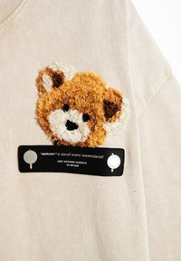 Sudadera beis con un aplique texturizado de cabeza de oso marrón y blanca, suave al tacto, con una etiqueta negra que dice "mod wave movement" y acentos plateados.