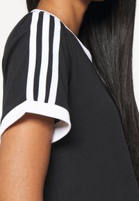 Černé tričko s bílými manžetami na rukávech a třemi bílými pruhy podél ramene. Hladká tkanina, ležérní design.