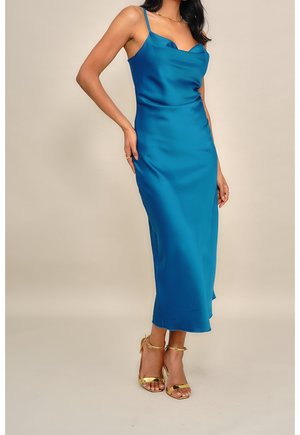 Femme portant une robe nuisette en satin bleu sarcelle avec bretelles spaghetti et des sandales dorées à talons hauts, debout devant un fond uni.