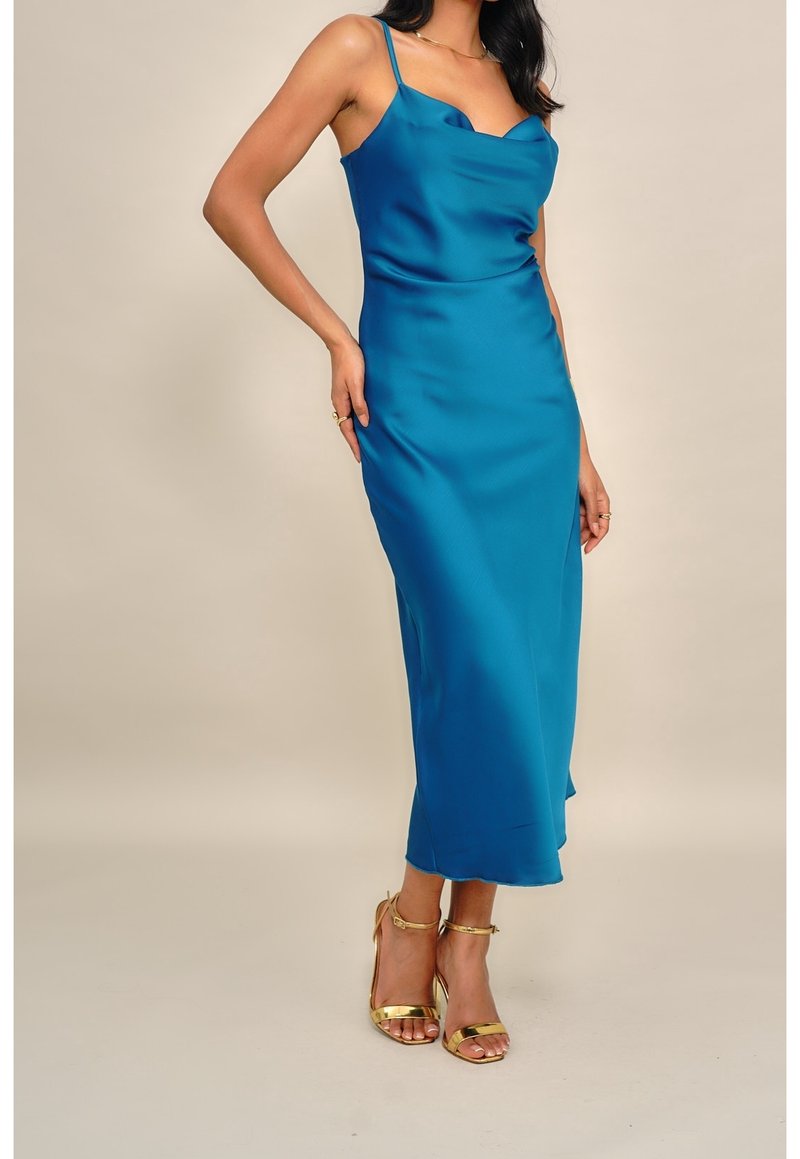 Femme portant une robe nuisette en satin bleu sarcelle avec bretelles spaghetti et des sandales dorées à talons hauts, debout devant un fond uni.