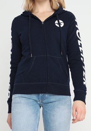Navy zip-up hoodie gemaakt van een katoenmix. Heeft een voorkant met rits, twee zijzakken, en witte tekst op de mouwen. Bevat een logo op de borst.