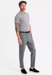 T-shirt grigio a maniche corte, pantaloni grigi slim-fit, scarpe slip-on nere, con tatuaggi ben visibili sugli avambracci. Sfondo semplice.