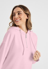 Lichtroze hoodie met een rits aan de voorkant, zilveren accenten en een ontspannen pasvorm. Glad materiaal en subtiele branding op de borst.