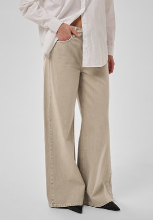 Person trägt beige Hosen mit weitem Bein, weißes Hemd mit Knöpfen und hat die Hand in der Tasche, schwarze spitze Schuhe auf schlichtem Hintergrund.