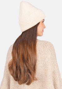 Bonnet en tricot côtelé couleur crème avec un sommet effilé, porté avec de longs cheveux bruns ondulés, sur un fond clair et uni.