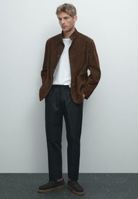 Hnědá nubuková bunda s knoflíkovým zapínáním, nošená přes bílé tričko. Spárováno s tmavými kalhotami a hnědými botami s gumovou podrážkou.