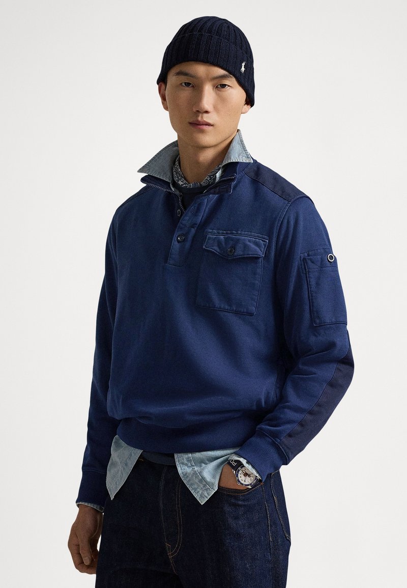 Temno modra pulover s ovratnikom, z zapenjanjem na gumbe, prsno žepom in kontrastnim teksturiranim materialom na ramenih. Nošena čez jeans srajco.