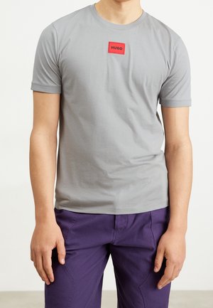 Hombre con camisa de manga corta gris claro con parche rojo "HUGO" y pantalones morados oscuros, de pie con los brazos relajados a los lados.