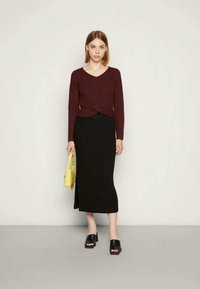 Jovem mulher vestindo uma blusa de manga longa burgundy, uma saia midi preta, sapatos de salto abertos pretos, segurando uma pequena bolsa amarela, em pé contra uma parede lisa.