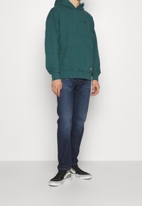 Turkusowa bluza z kangurzym kieszeniem i logo, zestawiona z ciemnoniebieskimi dżinsami oraz czarnymi trampkami z białymi podeszwami i szarymi akcentami.