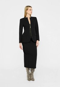 Zwarte, op maat gemaakte blazer met ritssluiting aan de voorkant, gecombineerd met een nauwsluitende zwarte rok en kniehoge laarzen met slangpatroon. Over het geheel een gladde textuur.