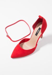 Escarpin à bout pointu en suède rouge avec bride fine amovible à la cheville et talon stilettos bas sur fond blanc.