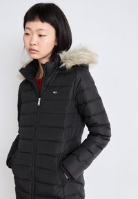 Mustast puffer-jakist, millel on esipoolsel külje tõmblukk, horisontaalne õmblustik, küljetaskud ja eemaldatav karvaga vooderdatud kapuuts. Logo vasakul rinnal.