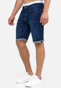 Shorts de mezclilla azul oscuro con puños enrollados, bolsillos frontales y cierre de botón. Llevados con zapatillas blancas para un look casual.
