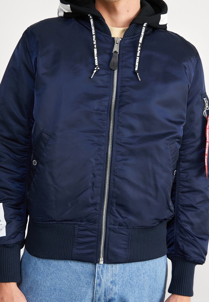 Homme portant une veste bomber bleu marine avec un sweat à capuche noir en dessous et un jean bleu clair, fermeture éclair et cordons visibles.