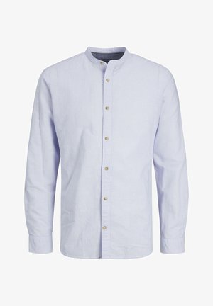Persona vestida con una camisa de botones azul claro, pantalones azul marino oscuro y zapatillas blancas, de pie contra un fondo claro y liso.