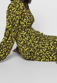 Černé květinové šaty se žlutými květinami, dlouhými nafouklými rukávy, hlubokým výstřihem a volnou sukní. Tkanina vypadá lehce a texturovaně.