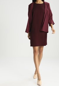 Vínová šaty s texturovanou látkou a odpovídajícím blejzrem. Blejzr má ohrnuté rukávy a strukturovaný design, doplněný o světle béžové lodičky.