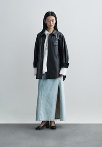 Zwart leren oversized overhemd met een zak, gecombineerd met een lichte denim maxi-rok met een zijsplit. Zwarte schoenen met blokhak.