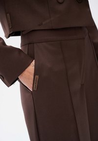Brązowa skórzana marynarka cropped z podwójnymi guzikami, uzupełniona dopasowanymi spodniami z bocznymi kieszeniami i metkę. Gładka faktura materiału.