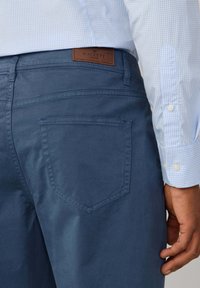 Granatowe bawełniane spodnie mają tylną kieszeń o teksturowanej powierzchni oraz brązową skórzaną łatę z napisem "HACKETT". Widać jasnoniebieską koszulę w kratę.