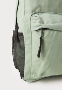 Mochila verde claro con tejido texturizado, acentuada por un bolsillo lateral de malla negra y un tirador de cremallera en contraste en la parte frontal.