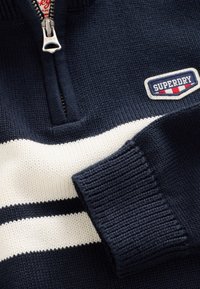 Námornícky modrý pulóver s bielymi horizontálnymi pruhmi, vybavený golierom so zipsom a textúrovanou pleteninou. Na hrudi je nášivka značky.