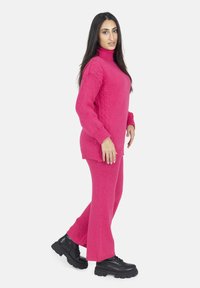 Helles pinkes Strickpullover mit hohem Kragen und Zopfmuster, kombiniert mit passenden Schlaghosen und klobigen schwarzen Stiefeln.