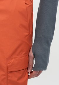 Tuvo plāksne ar roku, daļēji pārklātu ar pelēku piedurkni, blakus oranžai kabatām ar apģērbu.