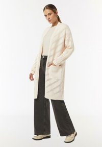 Langer Strickcardigan in hellbeige mit offener Vorderseite, strukturiertem Muster und diagonalem Streifen. Kombiniert mit grauen weit geschnittenen Hosen und beigen Schuhen.