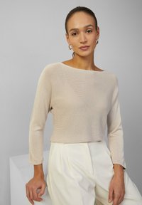 Hellbeige gestricktes Oberteil mit langen Ärmeln und weitem Ausschnitt, kombiniert mit weißen hoch-taillierten Hosen. Glatte Textur, lässiges Design.